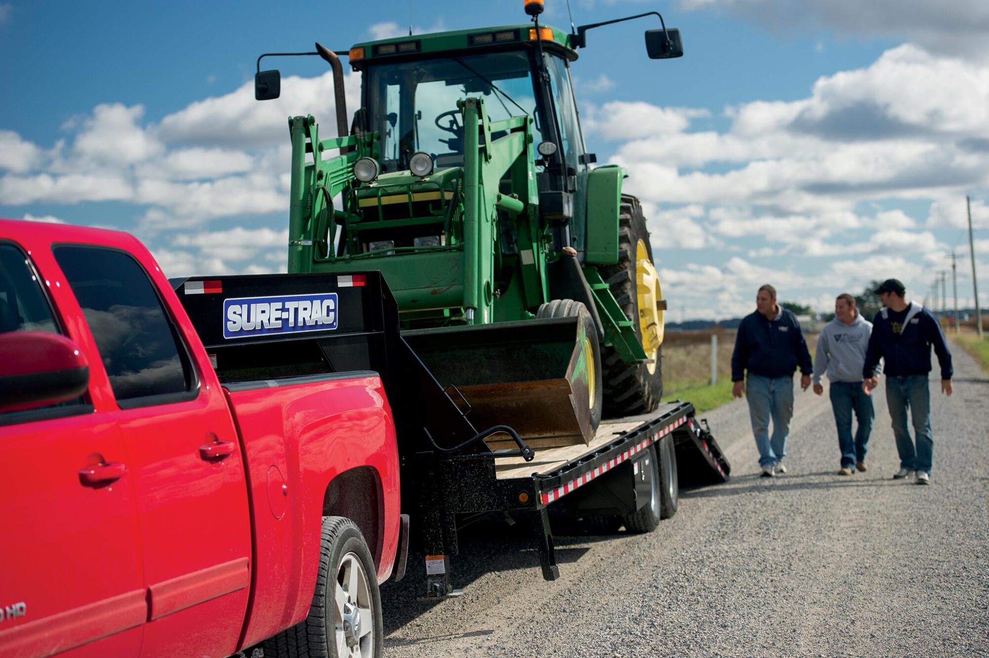 angled front view of the a tractor being pulled by a red truck on the heavy duty low profile hydraulic beavertail deckover trailer