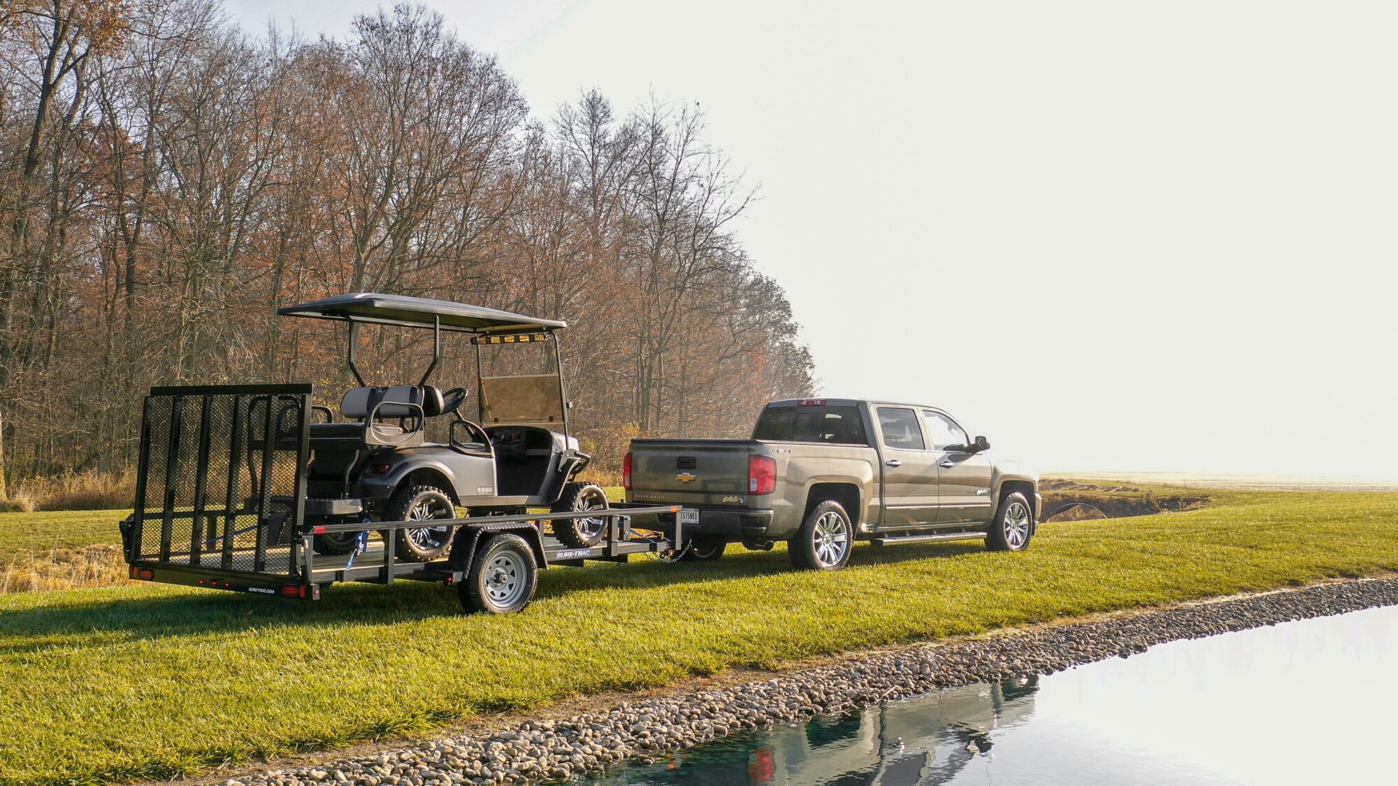truck pulling a trailer holding a golf cart next to an edge of trees
