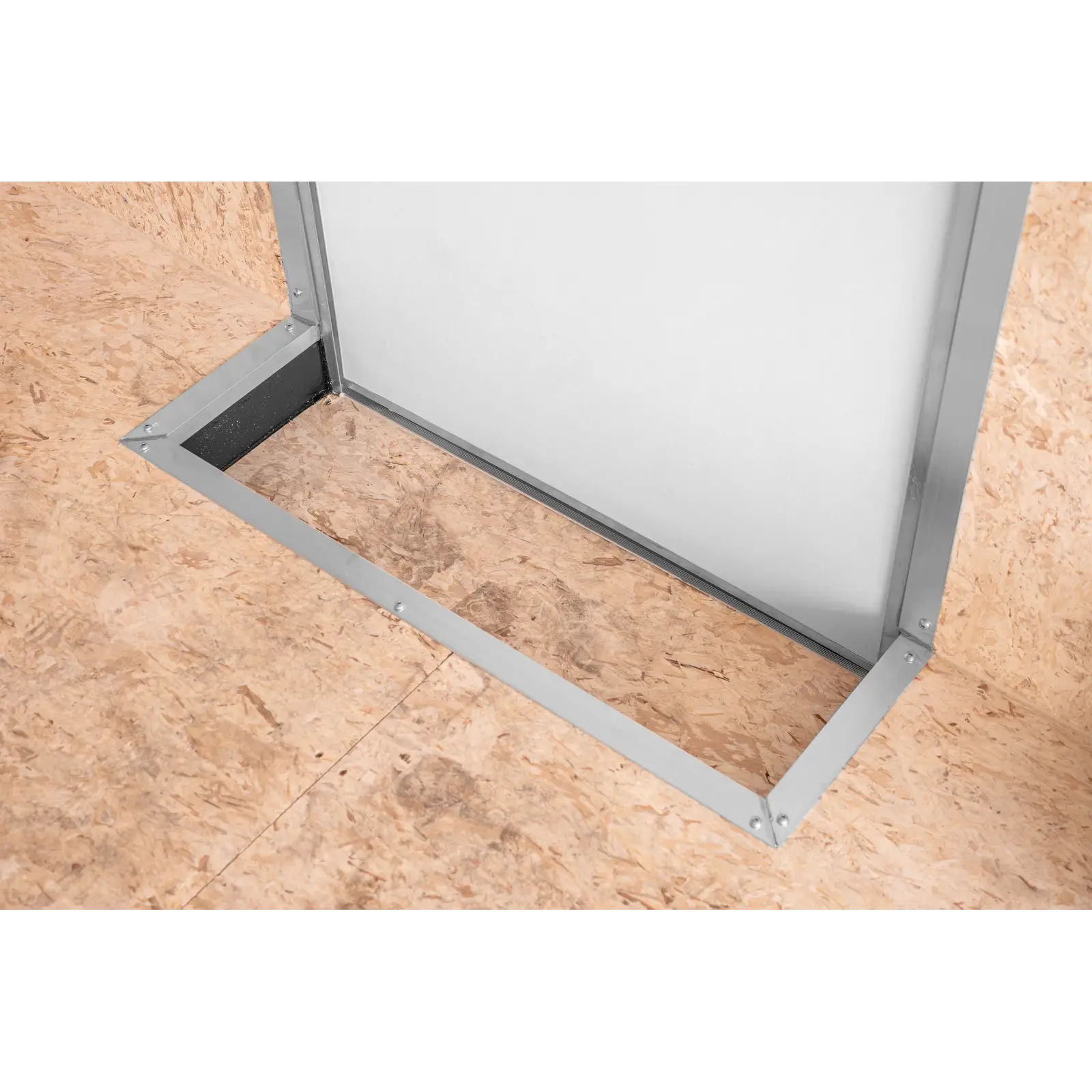 A top-down view of a square metal-framed hatch set in plywood flooring, with a white panel inside the frame and visible screws along the edges