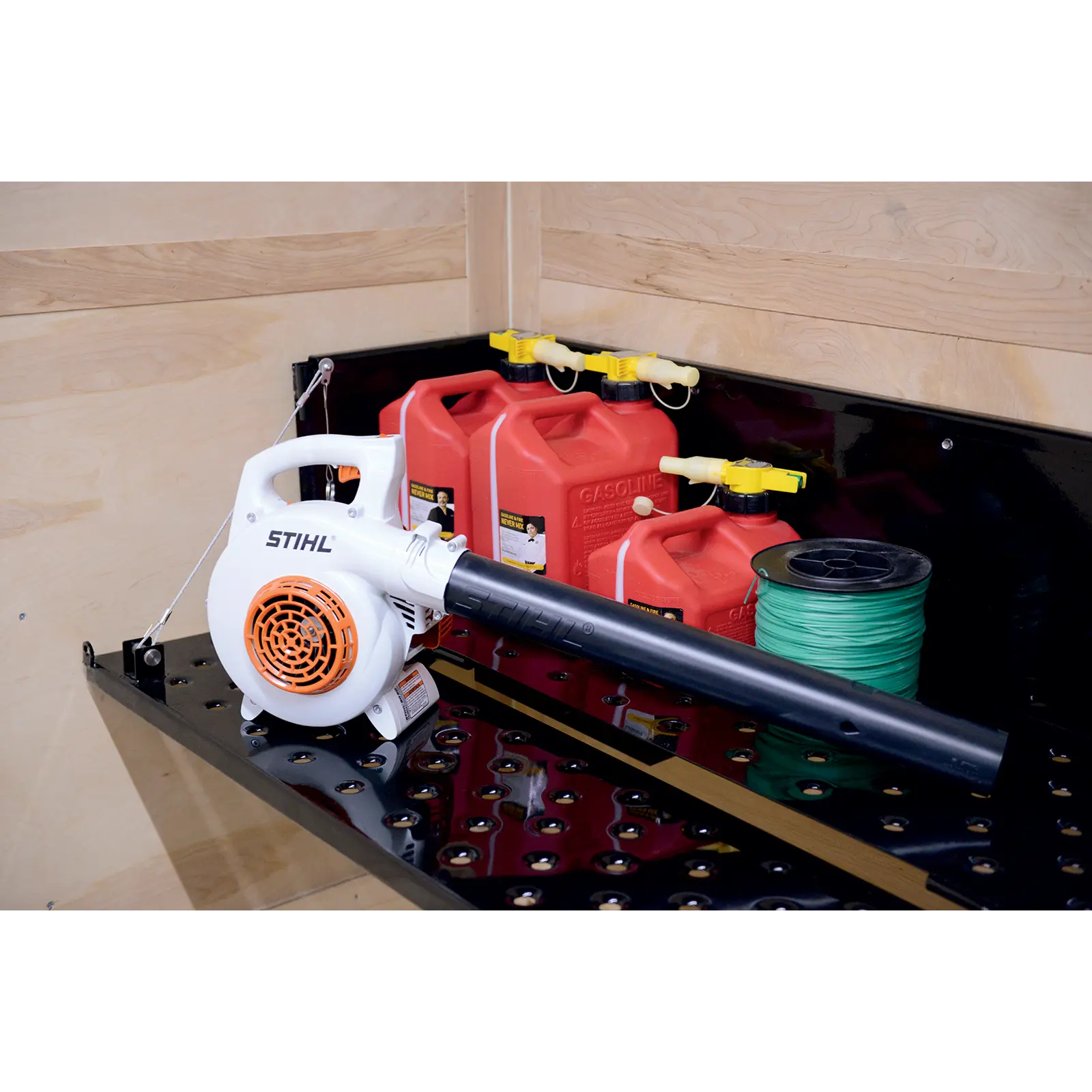 White STIHL leaf blower in a storage case with red gas cans and a roll of green spool on the right.